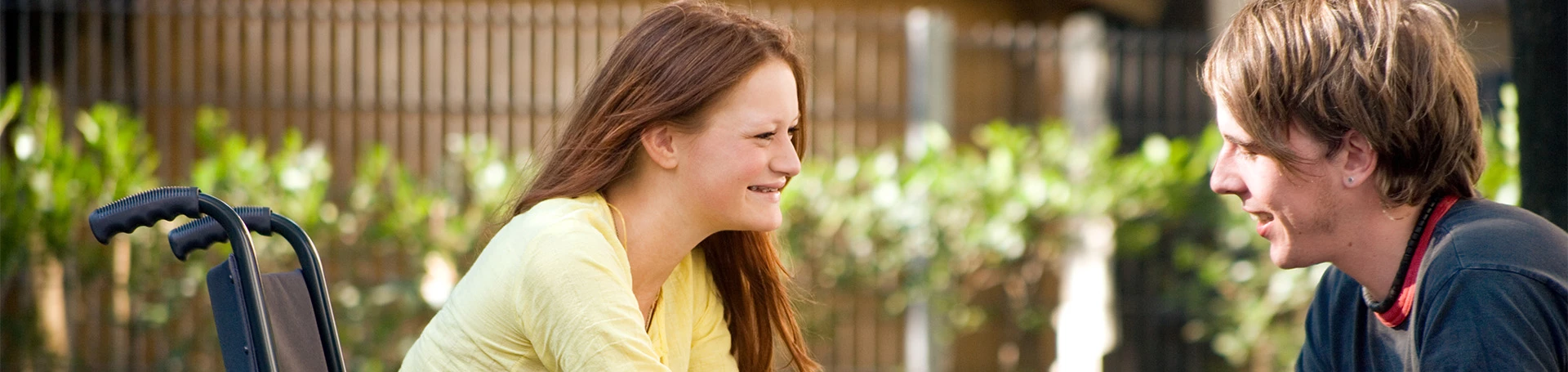 Chłopak i dziewczyna rozmawiają i uśmiechają się do siebie. Dziewczyna siedzi na wózku inwalidzkim.